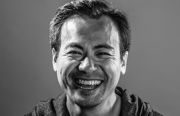 A smiling man with short dark hair looks directly at the camera in a black and white close-up portrait.