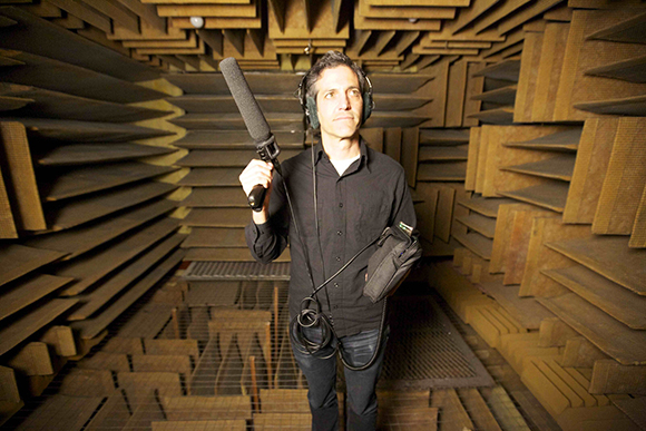 A man wearing headphones holds a large microphone inside a room lined with acoustic foam wedges.