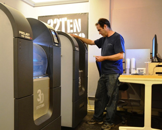 A man in a blue shirt stands beside large grey 3D printing machines in a workshop.