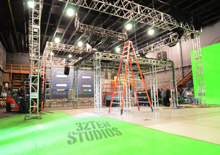 A film studio featuring metal scaffolding, overhead lighting rigs, a green screen, and a forklift.