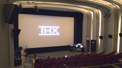 A large cinema screen shows a bright white THX logo in a dark theater with rows of red seats.
