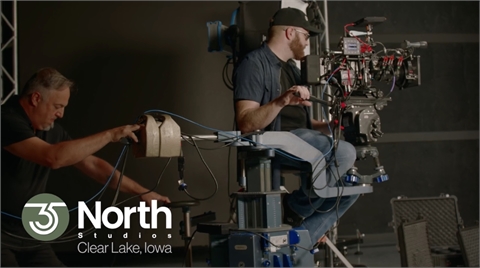 A man sits behind a large professional camera mounted on a mechanical arm in a dark studio.