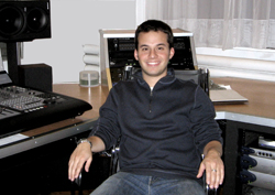 A smiling man in a dark zip-up sweater sits at a desk in a recording studio with audio equipment.