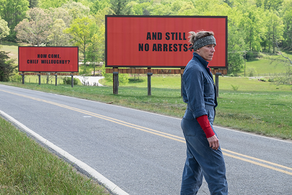 A woman in blue overalls and a headband walks along a road past two large red billboards.