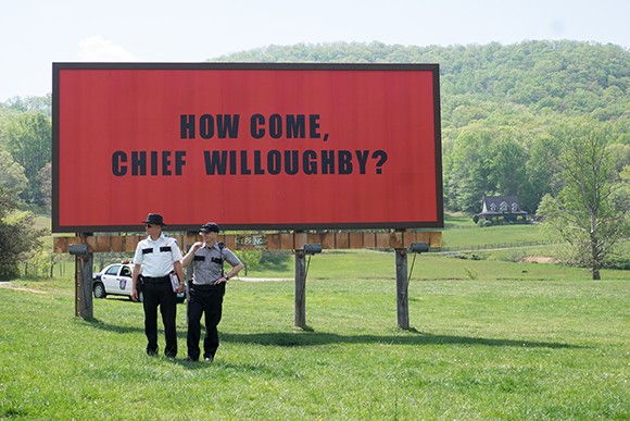 Two police officers stand in a grassy field before a large red billboard reading 'HOW COME, CHIEF WILLOUGHBY?'