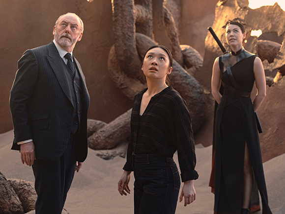 An older man in a suit and two women stand in a desert landscape with large, organic-looking rock formations.