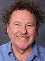 A smiling man with curly brown hair and a light beard wearing a blue collared shirt.