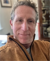 A close-up portrait of a man with curly hair wearing a brown fleece jacket.