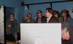 Several people wearing 3D glasses stand in a room, looking towards a large white screen. One person in the foreground is part