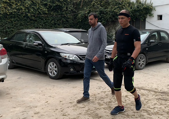 Two men walk past several parked cars in a dirt lot.