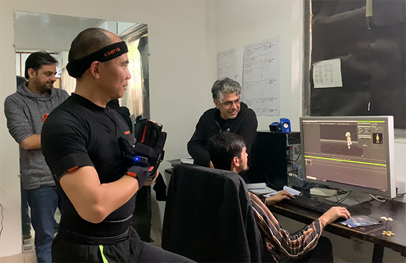 A man in a black motion capture suit and head sensor stands near technicians working at a computer.
