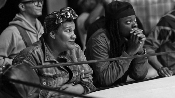 Two women sit behind a boxing ring rope, looking upward with focused expressions.
