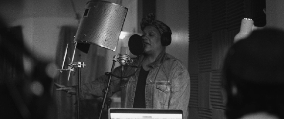 A person wearing headphones and a headwrap sings into a microphone in a recording studio.