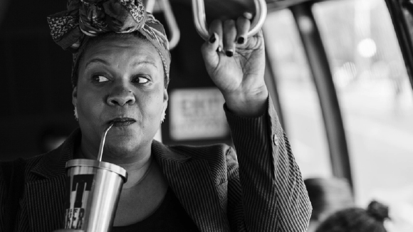 Black woman in a headwrap drinking from a metal cup while holding a handrail on a bus.