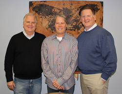 Three men standing side by side in front of a large piece of patterned wood art.