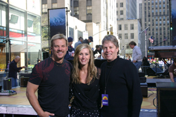 A man and a woman smile in the foreground with another man standing behind them outdoors.