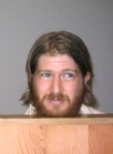 A man with long brown hair and a full beard looking toward the right against a gray background.