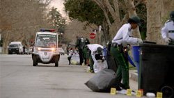 Investigators in white shirts and green pants examine evidence near trash bins on a street with a police vehicle.