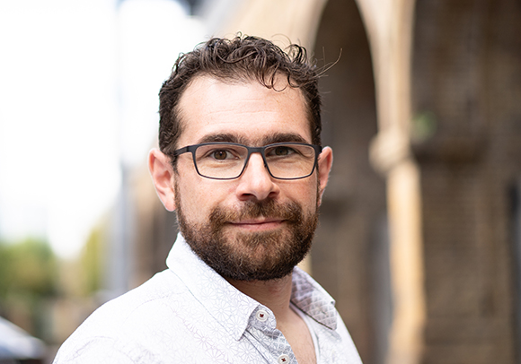 A man with dark curly hair, glasses, and a beard smiling against a blurred outdoor background.
