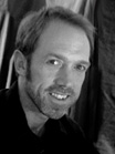 Black and white close-up portrait of a man with short hair and a light beard smiling.