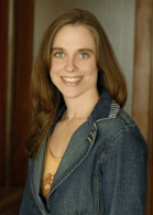 A smiling woman with long brown hair wearing a blue denim jacket over an orange top.