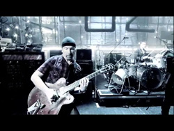 A man in a beanie plays an electric guitar in a studio with a drum kit behind him.