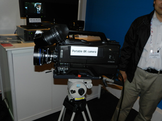 A portable 8K camera with a large lens is mounted on a tripod. A person stands to the right.