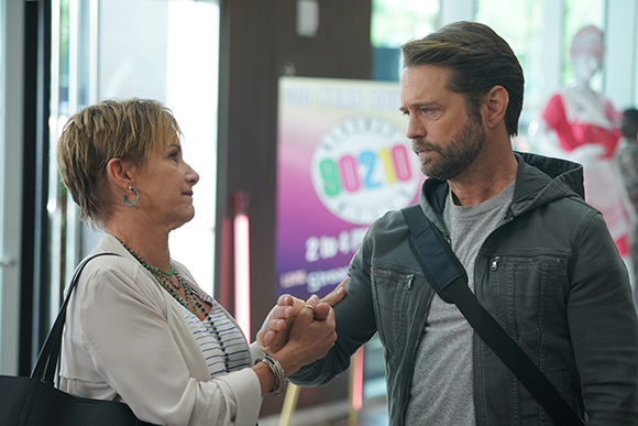 A woman with short blonde hair and a man in a grey hoodie hold hands and look at one another.