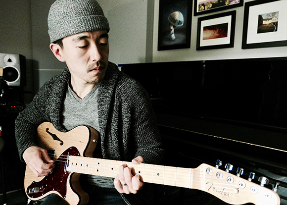 A man in a grey beanie and cardigan plays a light wood Fender electric guitar.