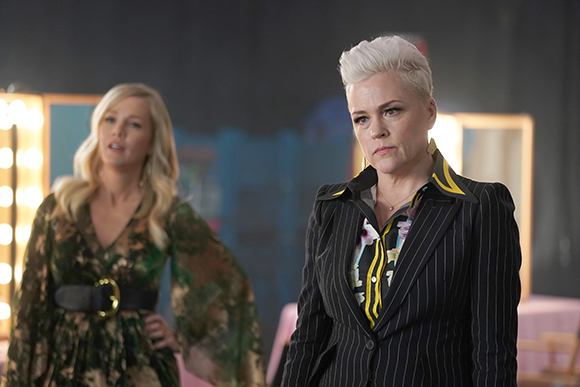 A woman with short white hair in a pinstripe blazer looks forward while another woman stands behind her.