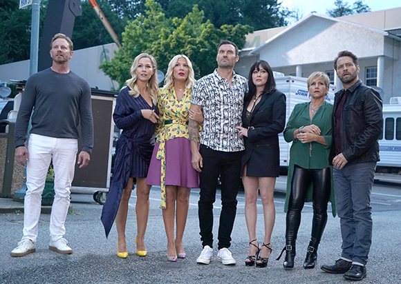 Seven people in various casual and semi-formal outfits stand in a line outdoors against a building and trees.