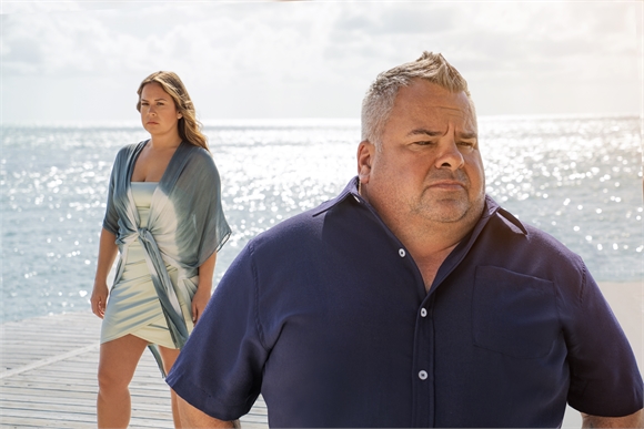 A man in a blue shirt looks forward while a woman in a blue dress stands behind him on a sunny pier.