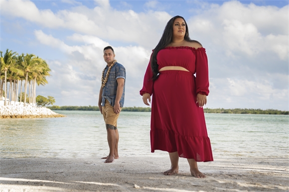 A woman in a red dress and a man in a blue shirt stand on a sandy beach under a cloudy sky.
