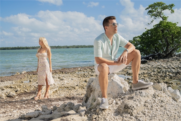 A man in a light shirt sits on rocks while a woman in a floral dress stands by the water's edge.