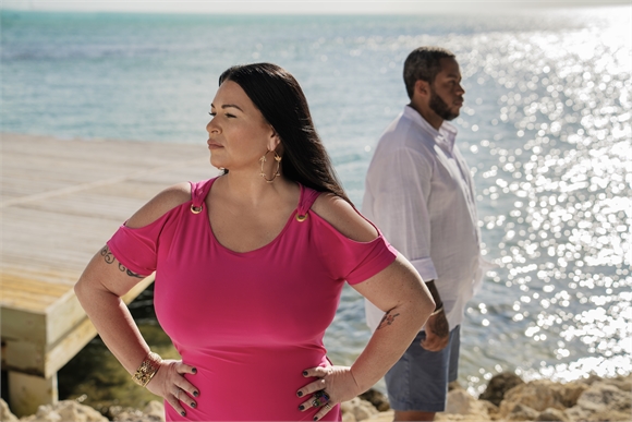A woman in a pink dress stands with hands on hips while a man in a white shirt stands behind her by the sea.