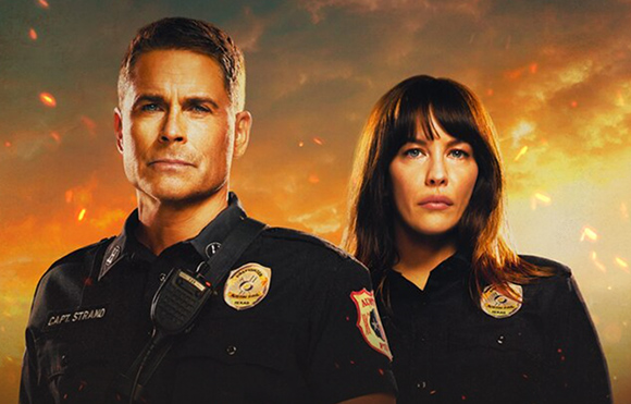 A man and a woman in dark firefighter uniforms stand before a backdrop of orange flames and smoke.