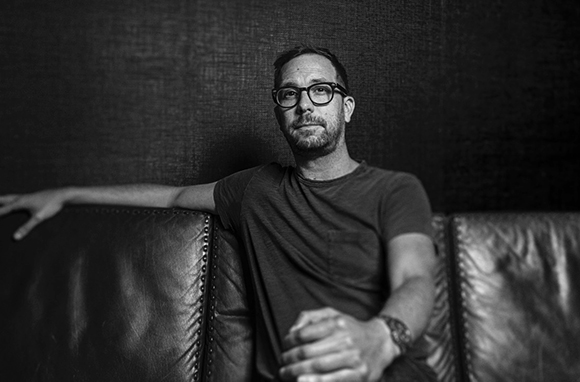 Black and white portrait of a man with glasses sitting on a leather couch against a dark wall.