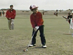 A young boy in a red shirt and dark pants swings a golf club on a grassy field.