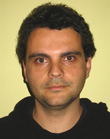 A man with dark curly hair and brown eyes looking directly at the camera against a yellow background.