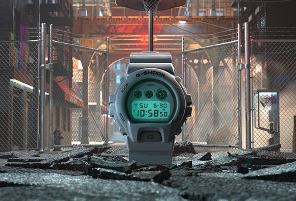 A grey digital G-Shock watch stands upright on broken pavement in a city alleyway with chain-link fences.