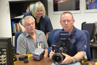 A woman and two men look closely at a large professional camera on a desk in an office.