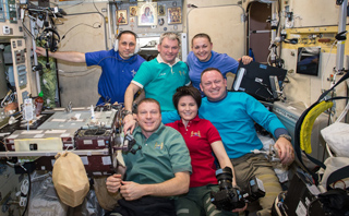 Six smiling astronauts in colorful shirts pose together inside a crowded space station module.