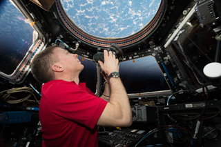 An astronaut in a red shirt looks through a circular porthole at Earth from a spacecraft.