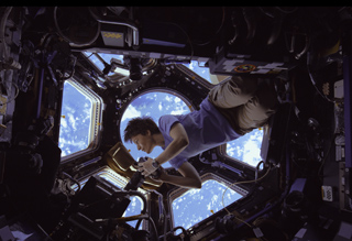 An astronaut floats in a dark spacecraft module, looking through a window at the blue Earth below.