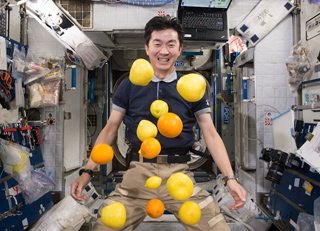 A smiling man in a dark polo shirt surrounded by floating lemons and oranges in a space station.