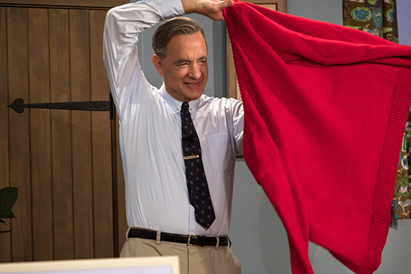 A man in a white shirt and tie holds up a large red sweater in front of a wooden door.