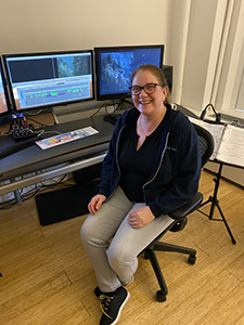 A smiling woman with glasses sits in an office chair at a desk with dual monitors.