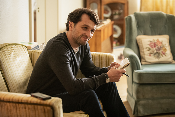 A man in a dark sweater sits on a striped sofa, looking ahead while holding a small notebook and pen.