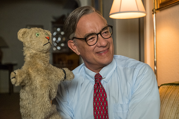 A man in a light blue shirt and red tie smiles warmly while looking at a worn teddy bear.
