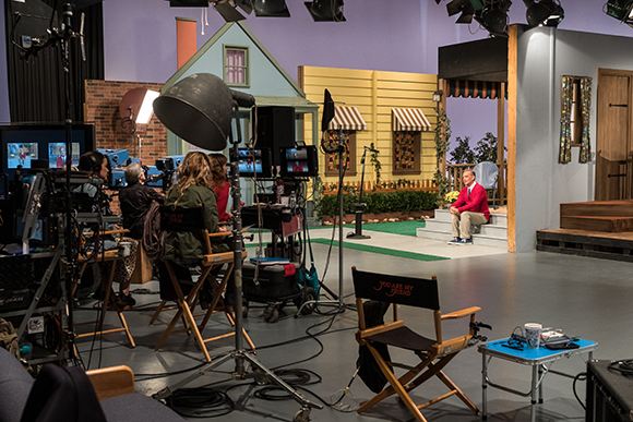 A television studio set with cameras, lighting equipment, and a man sitting on steps in front of a miniature house.